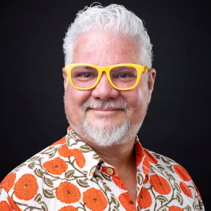 A man with short hair and a beard, wearing bright yellow glasses and a collared shirt patterned with large orange flowers and green leaves, smiles against a solid black background.