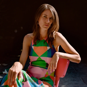 A poised woman in a vivid geometric-patterned dress sits in a red chair against a dark studio background, looking calmly toward the camera.