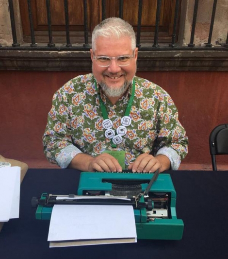 Image is of Leonardo Flores, a male person with tanned skin, white close-cut hair, and metal rimmed glasses. He is wearing a necklace with 5 silver medallions, as well as a long sleeved shirt with green and reddish print. He is smiling at the camera and sitting in front of a green typewriter. 